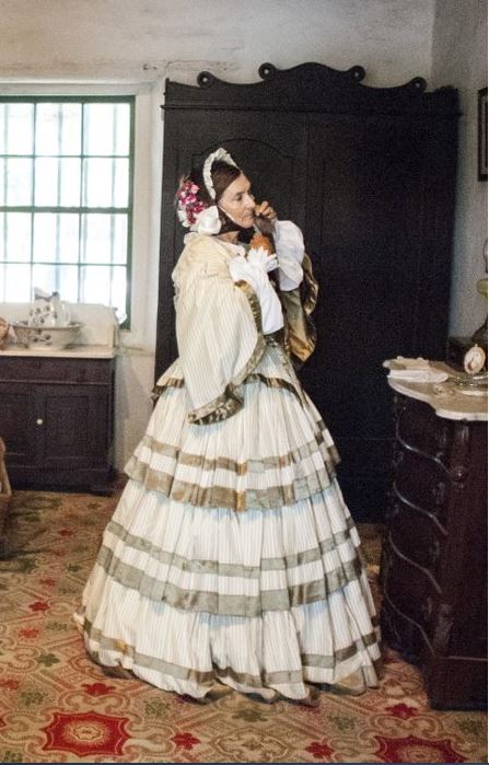 A woman in a long period dress looking in a mirror to tie her bonnet